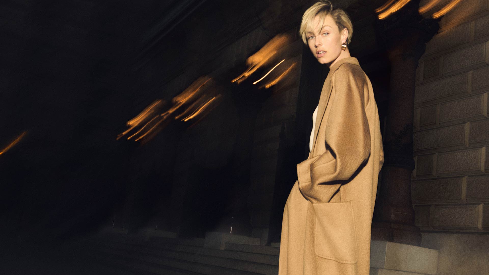 Model with Max Mara wide-fit Ludmilla icon coat in camel colour, in an evening setting.